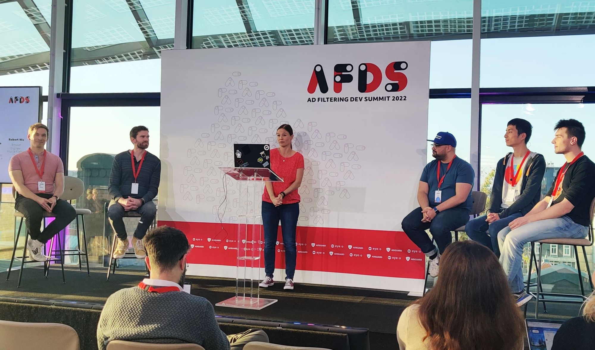 Six people on the stage, two sitting on high stools on the left, a moderator standing in the middle and three more sitting on high stools on the right