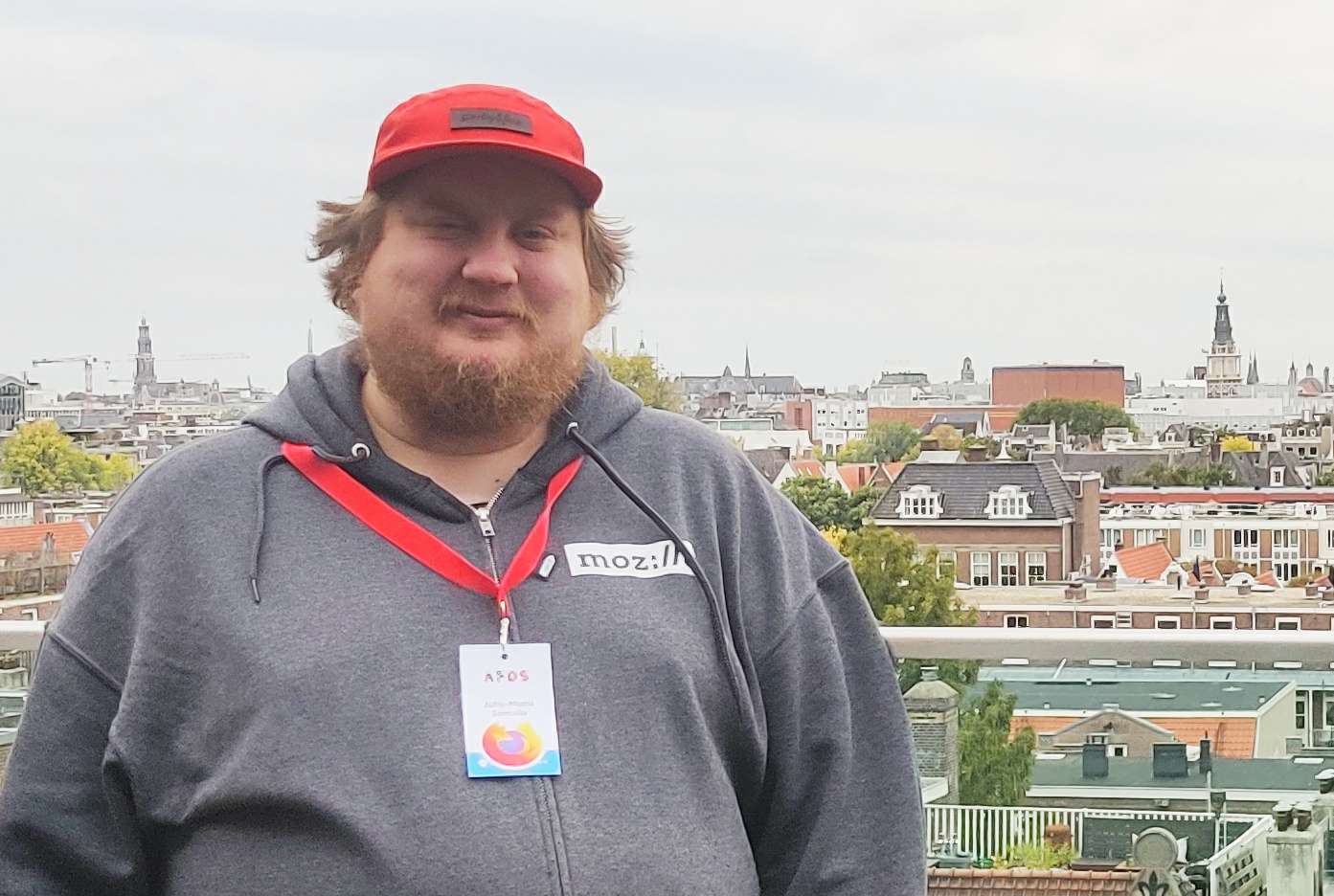 Me standing outside, wearing a red cap and grey hoodie with Mozilla logo. I have conference badge with a Firefox sticker hanging from my neck. Behind me, there are the rooftops of Amsterdam.