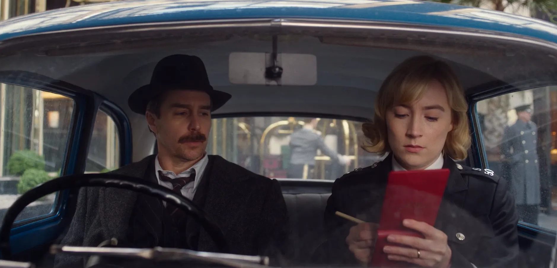 A man and a woman are sitting in a car, filmed from the front. The woman is writing notes into a red notebook and the man sitting next to her is looking at the notebook. 