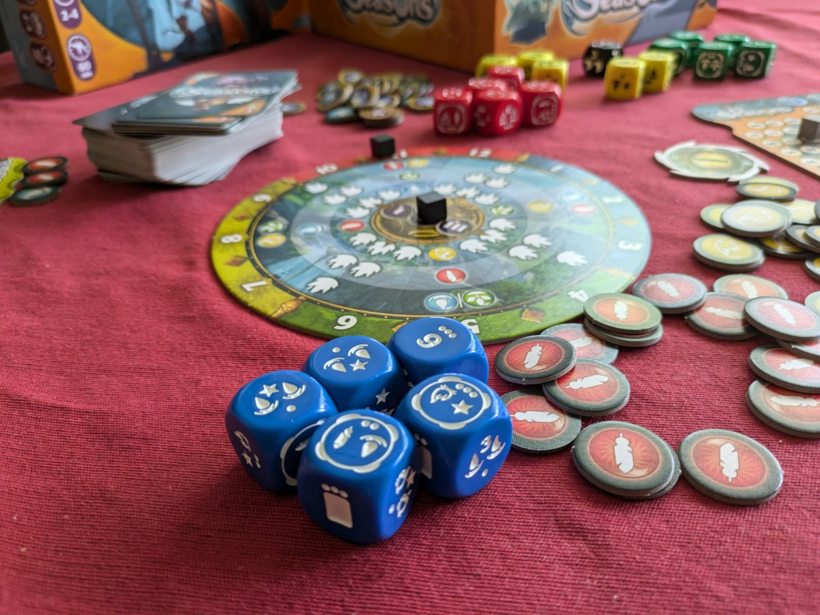 Bunch of board game components from Seasons spread on the table. In front, five blue dice with different markings, behind them a circle game board and scattered around some round energy tokens.