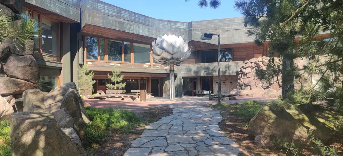 Outside of a two-story building with 60s architecture. In front of the building, basking in summer sun is a large metallic pine cone art installation that is surrounded by a small area for sitting around and a path made out of stone bricks leading into the yard. 