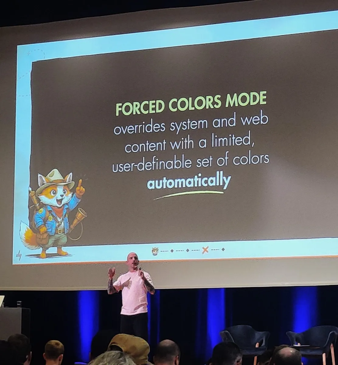 Daniel on the stage wearing a pink shirt. Behind him, a large screen with text: Forced colors mode overrides system and web contenr with a limited, user-definable set of colors automatically. 
