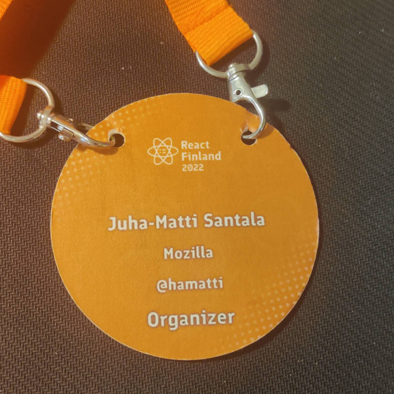 A round, bright orange conference badge for React Finland 2022 with the event logo, my name, company name, Twitter handle and role “Organizer”.