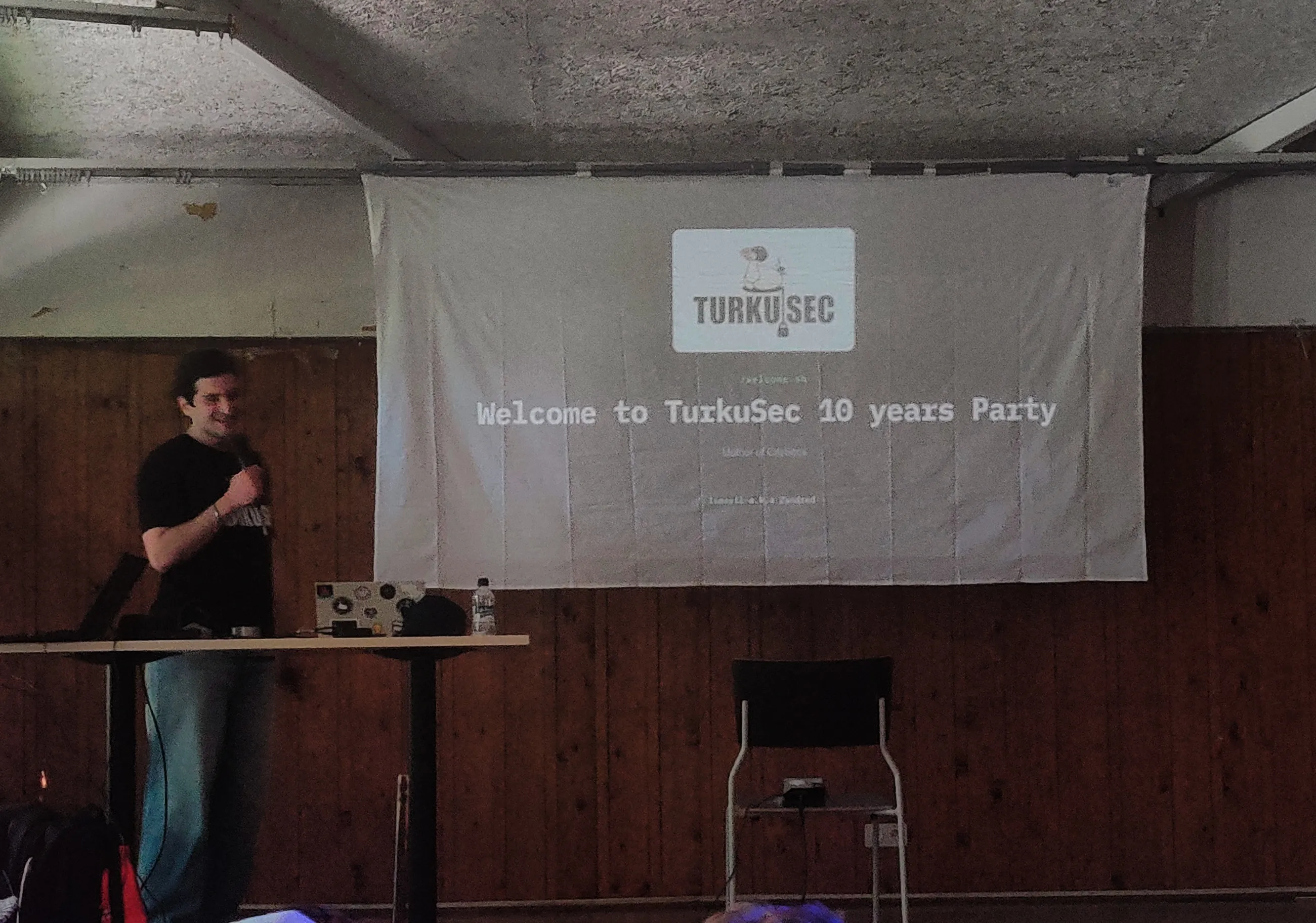 Ismayil standing next to a makeshift projector screen made of a bed linen. On it, a barely visible slide with title of Welcome to TurkuSec 10 years party 