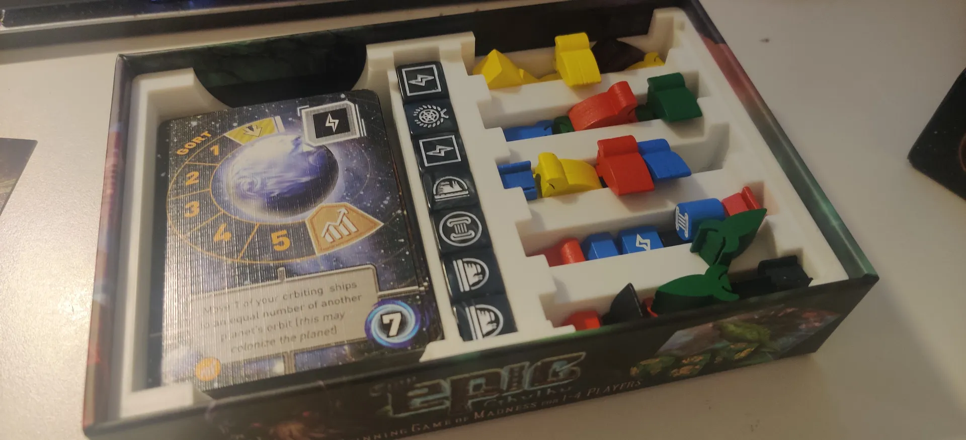 A Tiny Epic Galaxies board game box with a custom white 3D printed insert that has individual space for cards, dice and tokens. The tokens are bit all over the place and have been mixed up instead of staying in their crevices. 