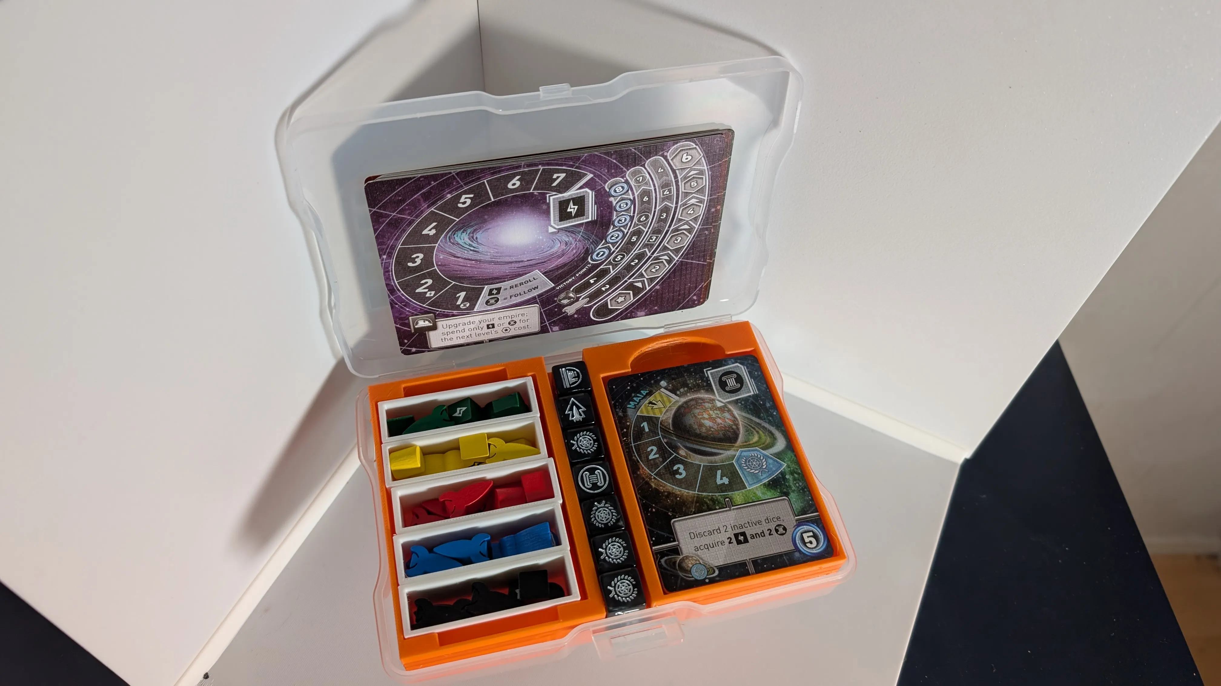 An orange 3D printed insert for Tiny Epic Galaxies inside a transparent plastic photo caddy box. On the left side area of the insert, there’s are 5 narrow white containers with tokens separated into them by five colours: green, yellow, red, blue and black. 