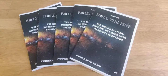 Five copies of the same zine fanned out on a table. The zine is Roll the Zine's July 2026 issue, a Finncon special: To boldly play where no one has played before. 