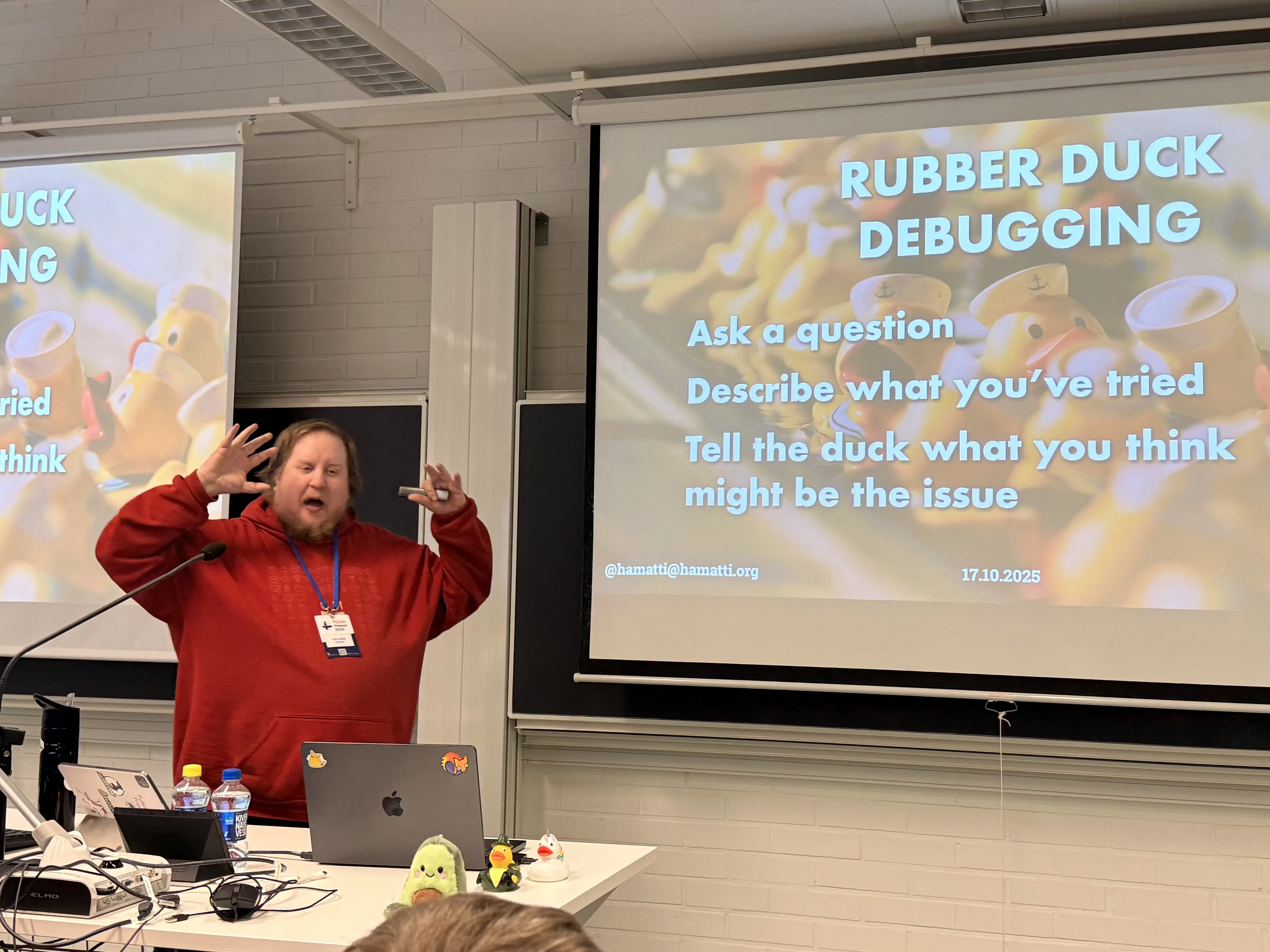 Juhis is standing in front of a room presenting with both of his arms up like he’s showing that something is very big. Next to him is a slide with a picture of yellow rubber ducks in sailor costumes and title Rubber duck debugging. Next to his laptop on the table there’s an avocado plushie and two rubber ducks. 