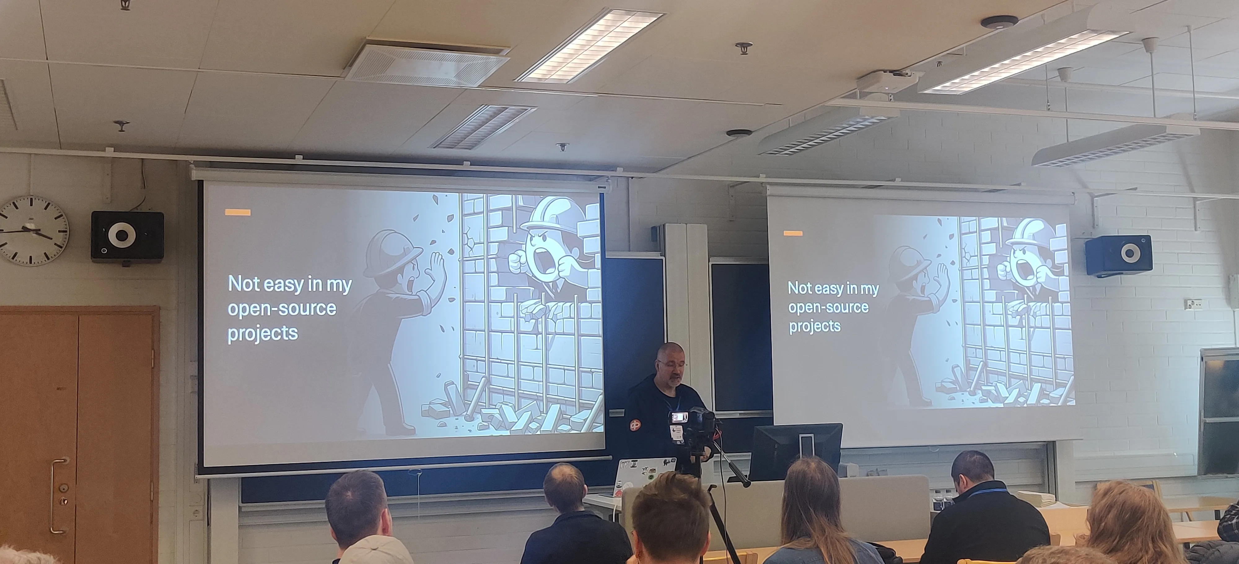 Tatu is presenting in front of a room with a slide that has an image of two construction workers yelling at each other through a broken wall and title Not easy in my open-source projects. 