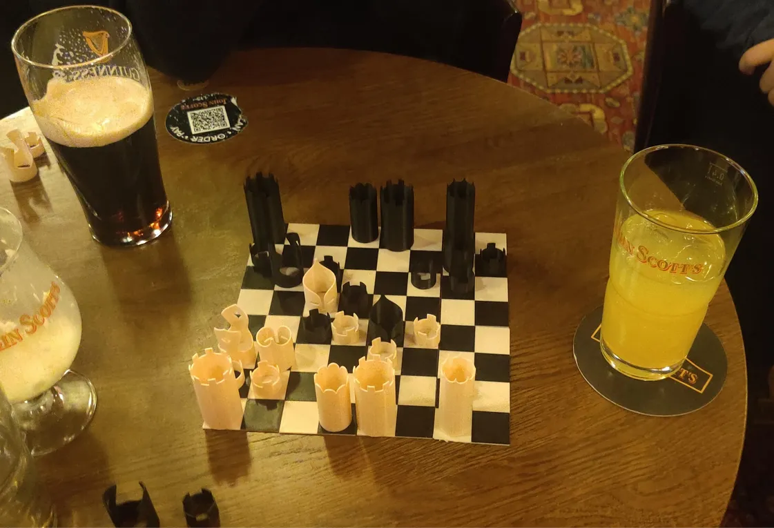 A small, 3D printed chess board with pieces being played in a pub, surrounded by drinks.