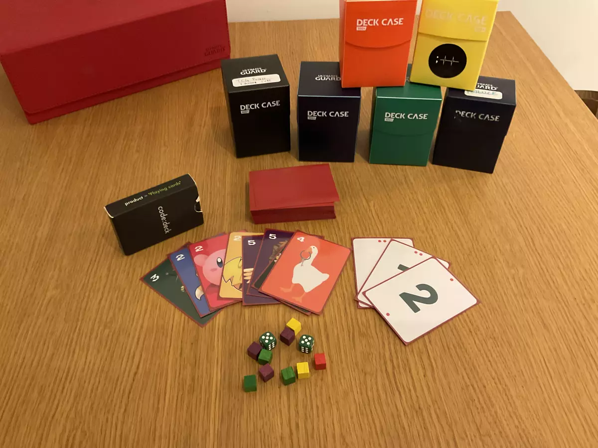 A bunch of deck boxes in various colors in the background and on the front, a deck of sleeved cards face down and a couple of colourful cards fanned out next to the deck. The cards have different vibrant background colours in orange, purple, yellow, red, blue and green and video game characters on them, the goose from Untitled Goose Game on the top most card.