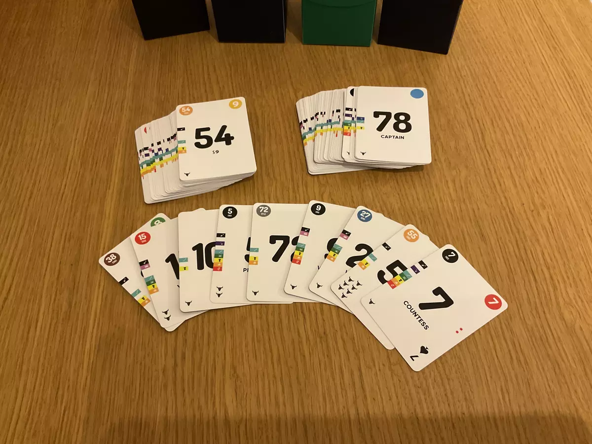 Two piles of cards with large numbers 54 and 78 on top-most cards. In front, a bunch of cards from the same decks fanned out showing different elements of the graphic design on the cards from different coloured numbers to traditional card suits and in the bottom left corner of each card, different amounts of small bullheads.