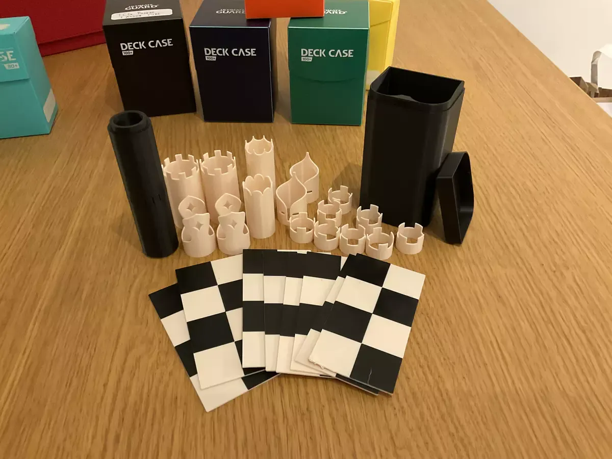A tiny chess set with a small black case. The black chess pieces are stacked up in a round tube inside each other and the white pieces are spread out showing their tubular design. In front of the pieces, 8 pieces of 4 times 2 chess board fanned out. 