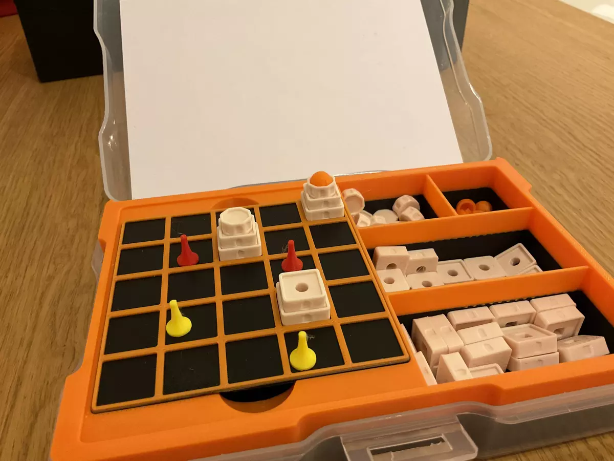 A tiny 5 by 5 black and orange playing board inside a clear plastic case and an insert that holds the board and has four smaller compartments for various game pieces. On the board, there are four player pieces in red and yellow as well as a couple of white buildings stacked from two to four pieces each with an orange dome on top of one of them.