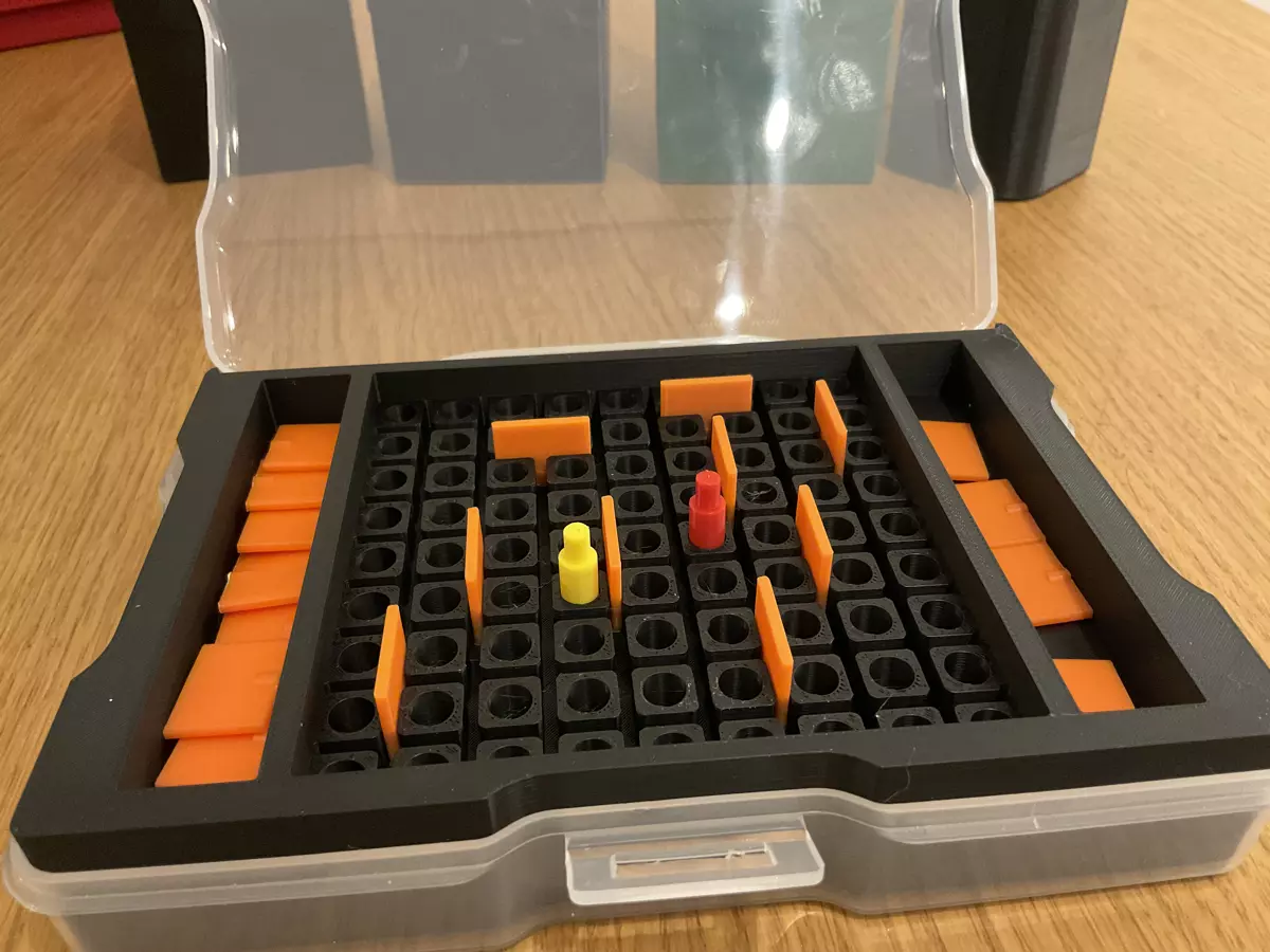 A black 3D printed insert inside a clear box. Inside the insert, there are 81 small circular holes and between them, space for tiny 3D printed orange walls. Some walls have been inserted into the game board with a red and yellow player meeples in two of the round holes.