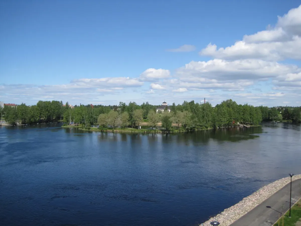 An ocean with a tree covered island on a beautiful sunny summer day. The island has a white church and lots of trees partially covering it from the view. The ocean is deep blue. 