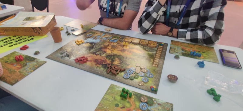 A game of Stone Age set up for four players with a main board, each player with their own player board and colored meeples and a lots of wood, clay, stone, gold and food tokens on the board.