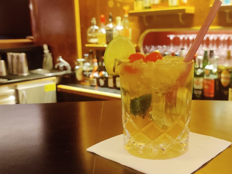 A colorful drink on a bar with the drink selection of the bar in the background out of focus.