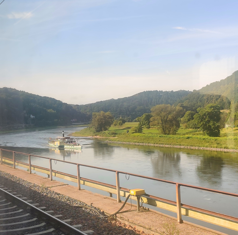A photo taken from the train with a river and a river boat and on the other side of the river, a patch of land with trees and hills rising from beyond that land