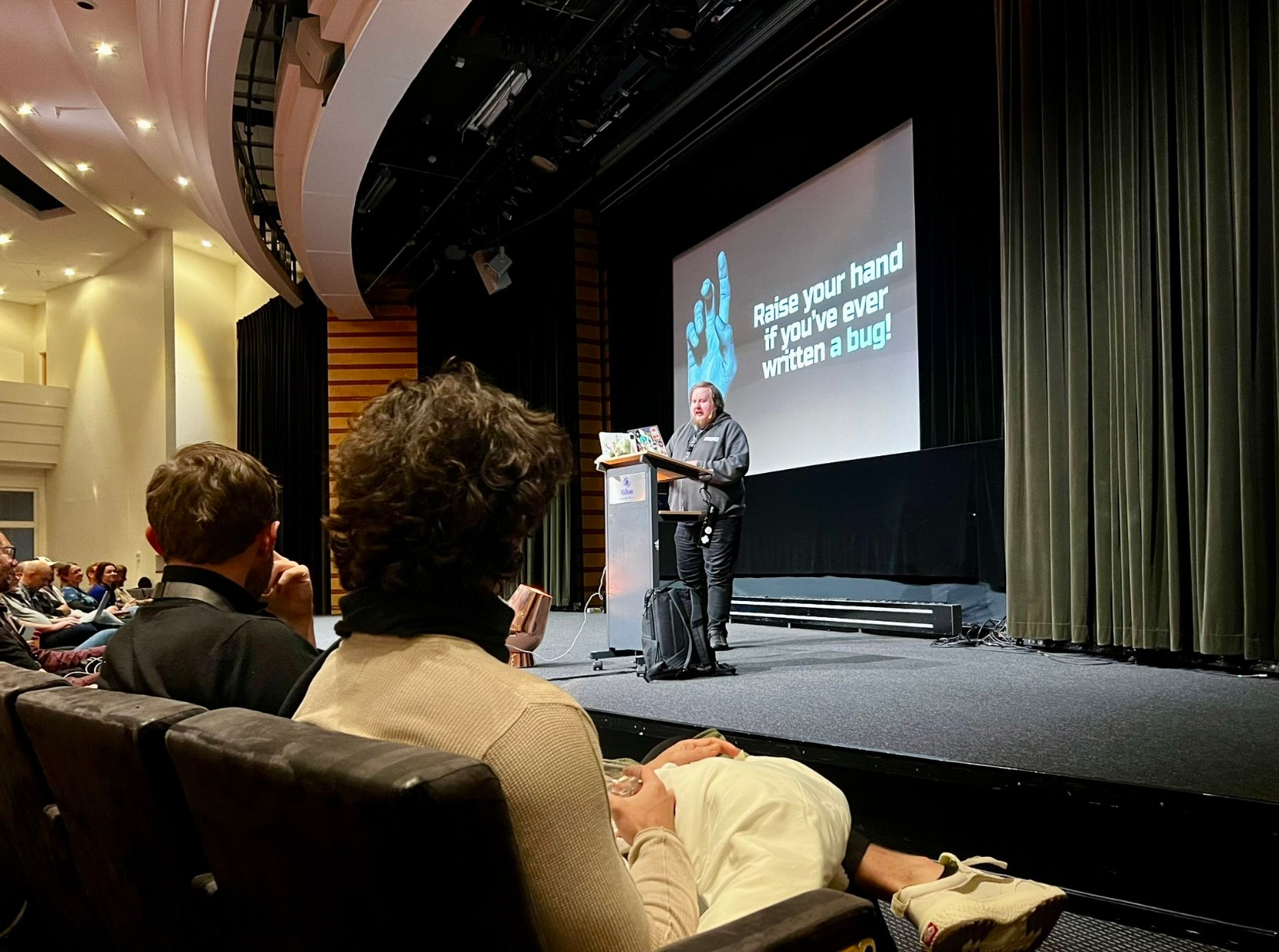 Juhis standing on stage next to a speaker podium. Behind him, a slide that has a raised hand and a text “Raise your hand if you’ve ever written a bug!”